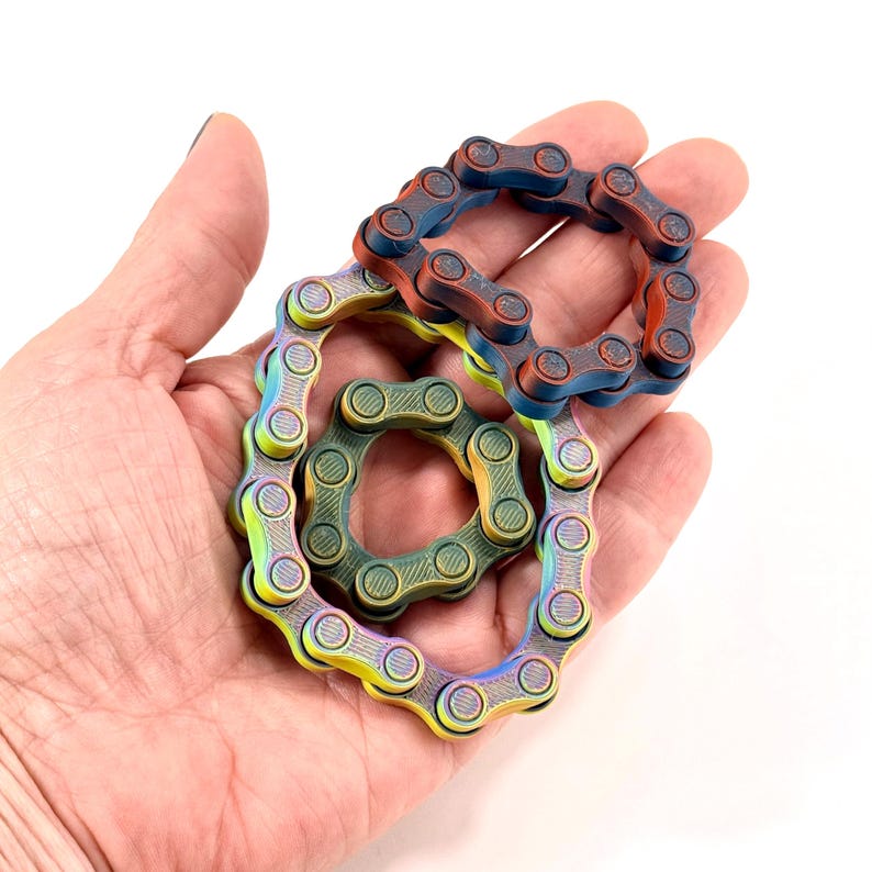 Puede incluir: Tres juguetes antiestr&eacute;s circulares construidos con eslabones de cadena de bicicleta de metal interconectados. Un anillo presenta un degradado azul y cobre, otro es verde oscuro s&oacute;lido y el tercero muestra un acabado arco&iacute;ris.