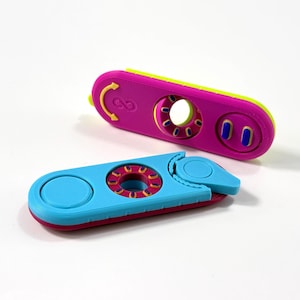 Könnte beinhalten: Zwei längliche Fidget-Spielzeuge mit taktilen Elementen. Das obere Spielzeug ist magentafarben mit einem lindgrünen Rand, einer kreisförmigen Aussparung, zwei blauen Knöpfen und einem Unendlichkeitssymbol. Das untere ist hellblau mit magentafarbener Basis.