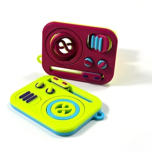 Könnte beinhalten: Zwei rechteckige Fidget-Spielzeuge mit abgerundeten Ecken. Eines ist magentafarben mit einem großen kreisförmigen Zentrum, drei vertikalen blauen Balken und zwei kleinen Knöpfen. Das andere ist limettengrün mit einem großen kreisförmigen Zentrum.