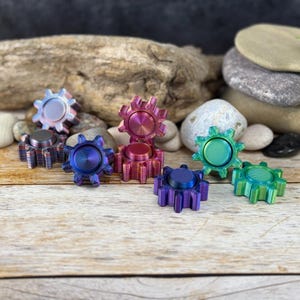 Può includere: Una collezione di fidget spinner a forma di ingranaggio colorati. Gli spinner presentano una gamma di tonalità, tra cui viola, blu, rosa e verde, con una lucentezza metallica. Sono disposti su una superficie di legno con uno sfondo di pietra naturale.