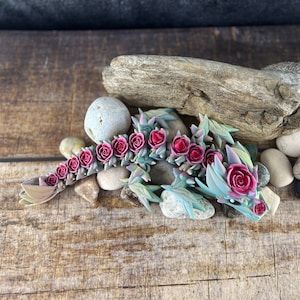 Può includere: Una piccola statuina di drago verde e iridescente con accenti di rose rosa. Il drago è sdraiato su una superficie di legno con pietre e legno galleggiante.