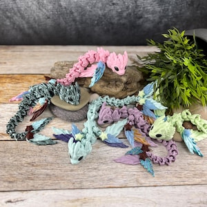 Può includere: Cinque piccole figurine di drago segmentate, colorate, con ali e dettagli iridescenti. I draghi sono disposti su una superficie di legno con una roccia e fogliame verde sullo sfondo.