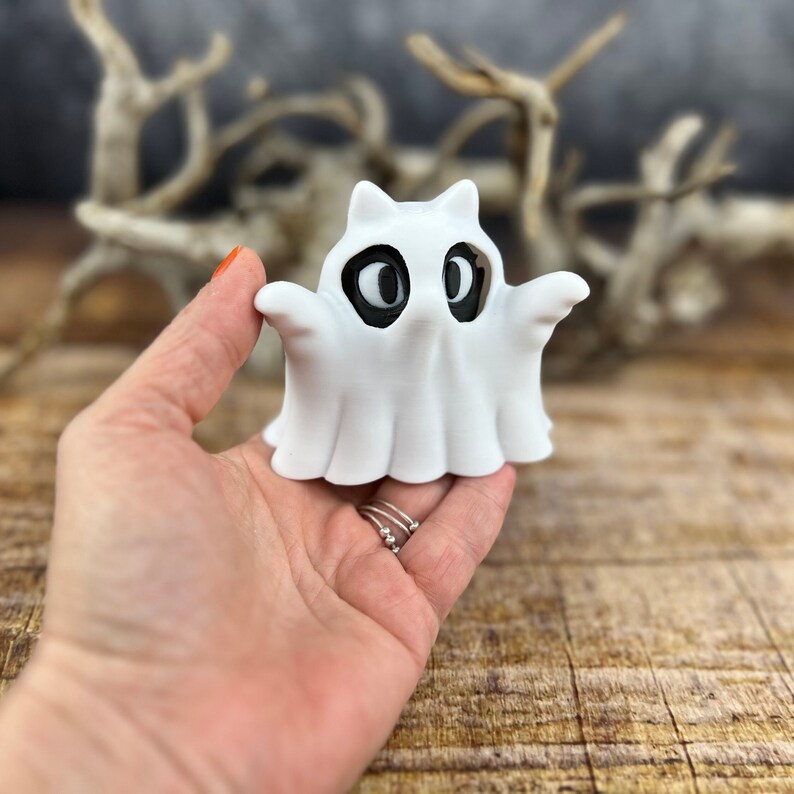 May include: White ghost figurine with black eyes. The ghost has a rounded shape and is holding its arms out.