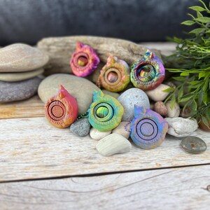 Peut inclure: Six jouets sensoriels en métal irisé en forme de flammes avec un centre rotatif. Les jouets sont disposés en cercle sur une surface en bois avec des pierres.