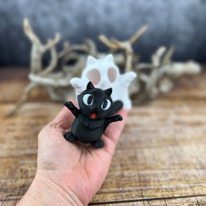 May include: A black cat figurine with big eyes and a wide smile. The cat is standing on a hand with a white ghost figurine behind it.