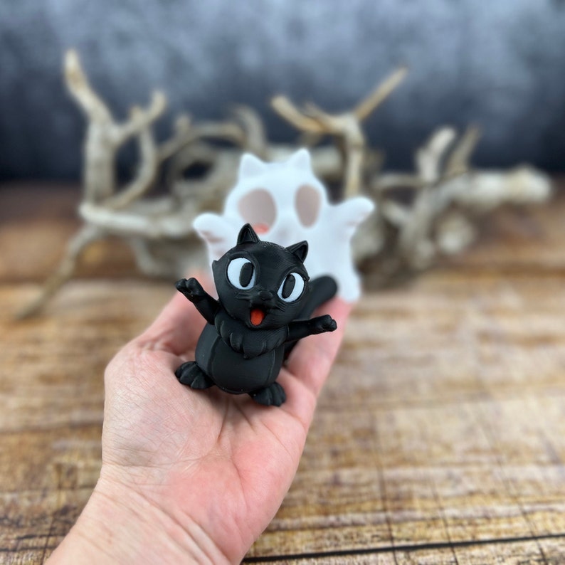 May include: A black cat figurine with big eyes and a wide smile. The cat is standing on a hand with a white ghost figurine behind it.