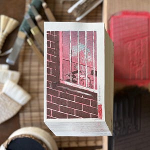 May include: A rectangular block of paper featuring a woodblock print of a brick wall and a window with vertical bars. The print uses shades of brown and pink, with the word Reflection and a small red stamp visible on the right side of the paper.