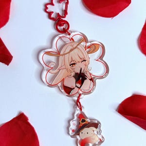 Puede incluir: Un charm de acrílico transparente con un personaje de anime de dibujos animados con cabello rubio y un guiño, sujeto a un cierre rojo en forma de flor. Debajo, un charm más pequeño representa un personaje con un sombrero de bruja. Acentos rojos y una cuerda roja.