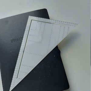 Puede incluir: Un cuaderno negro con la palabra "BULLET" en relieve en la portada, parcialmente oscurecido por una regla triangular blanca. La regla presenta medidas en centímetros y marcas fraccionarias, ideal para una maquetación y diseño precisos.