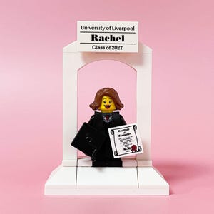 May include: A Lego figurine of a graduate in a black gown and mortarboard, holding a diploma and a cap. The figurine is standing in front of a white archway with a sign that reads "University of Liverpool, Rachel, Class of 2027" against a pink background.
