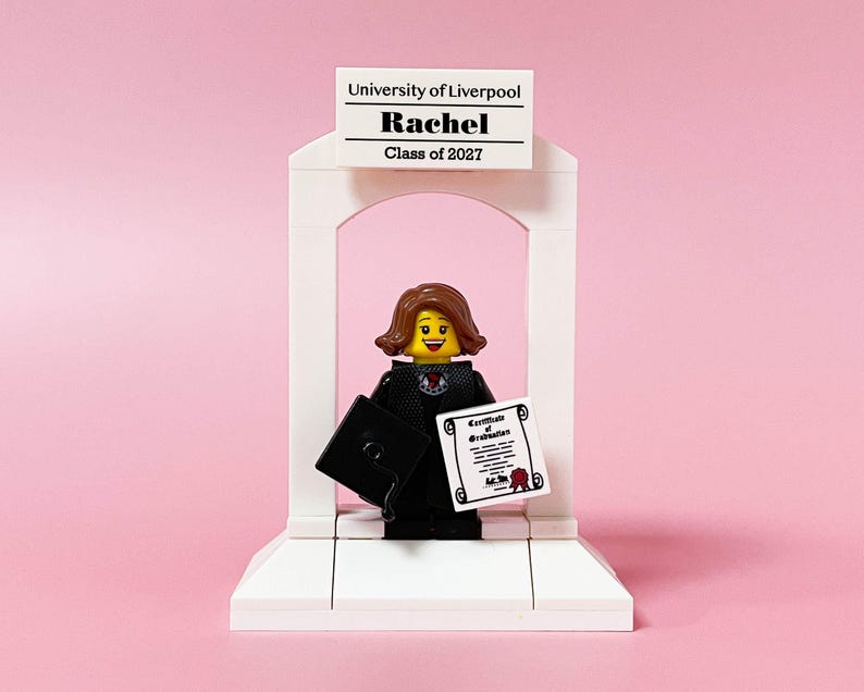 May include: A Lego figurine of a graduate in a black gown and mortarboard, holding a diploma and a cap. The figurine is standing in front of a white archway with a sign that reads "University of Liverpool, Rachel, Class of 2027" against a pink background.