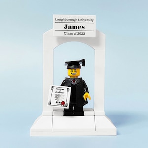 May include: A Lego figure wearing a black graduation gown and cap, holding a diploma, standing in front of a white archway with a sign that reads "Loughborough University James Class of 2023".