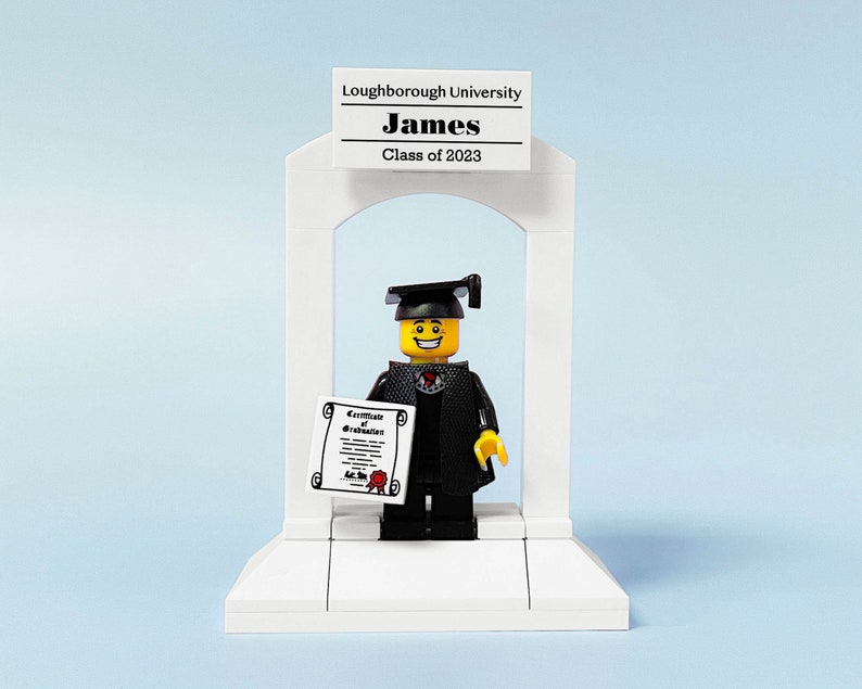May include: A Lego figure wearing a black graduation gown and cap, holding a diploma, standing in front of a white archway with a sign that reads "Loughborough University James Class of 2023".