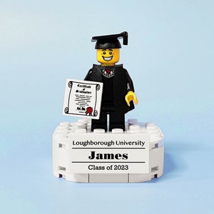 May include: A Lego figure wearing a black graduation gown and cap, holding a diploma. The figure is standing on a white platform with the text "Loughborough University, James, Class of 2023".