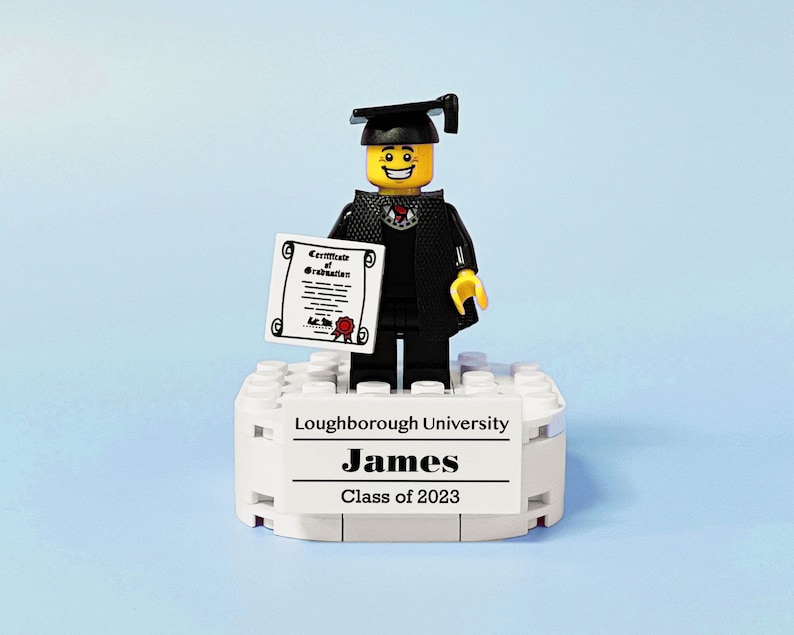 May include: A Lego figure wearing a black graduation gown and cap, holding a diploma. The figure is standing on a white platform with the text "Loughborough University, James, Class of 2023".