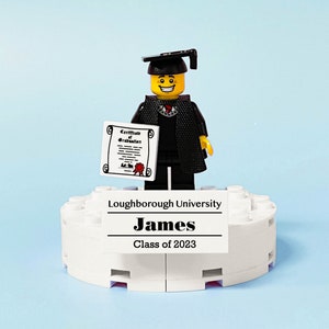 May include: A Lego figure wearing a black graduation gown and cap, holding a diploma. The figure is standing on a white platform with the text "Loughborough University", "James", and "Class of 2023".