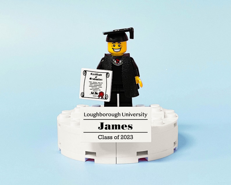 May include: A Lego figure wearing a black graduation gown and cap, holding a diploma. The figure is standing on a white platform with the text "Loughborough University", "James", and "Class of 2023".