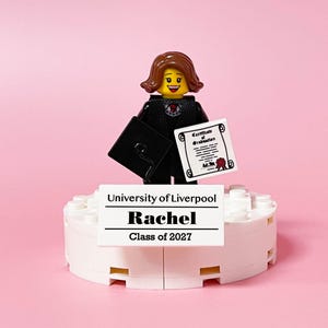 May include: A Lego figurine of a graduate in a black gown and mortarboard holding a diploma. The figurine stands on a white platform with the text "University of Liverpool, Rachel, Class of 2027" against a pink background.