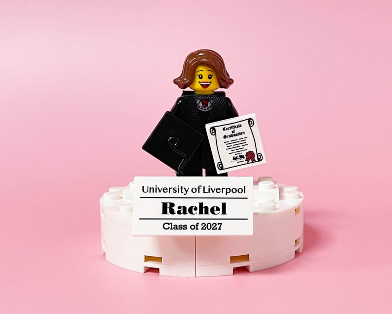 May include: A Lego figurine of a graduate in a black gown and mortarboard holding a diploma. The figurine stands on a white platform with the text "University of Liverpool, Rachel, Class of 2027" against a pink background.