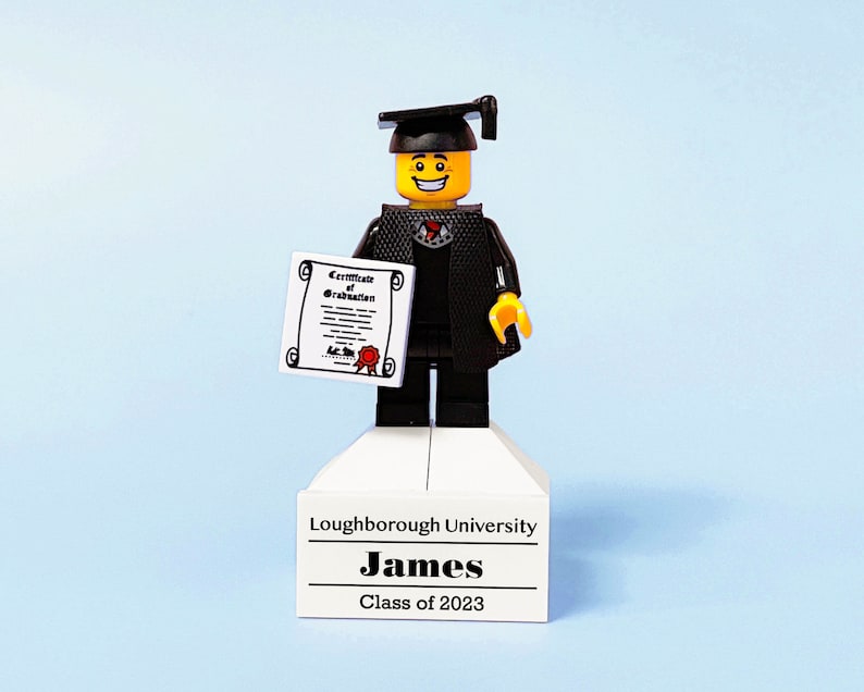May include: A Lego figure wearing a black graduation gown and cap, holding a diploma. The figure is standing on a white platform with the text "Loughborough University, James, Class of 2023".