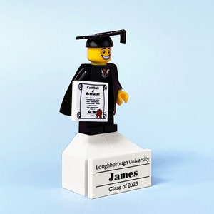 May include: A Lego figure wearing a black graduation gown and cap, holding a certificate. The figure is standing on a white platform with the text "Loughborough University, James, Class of 2023".