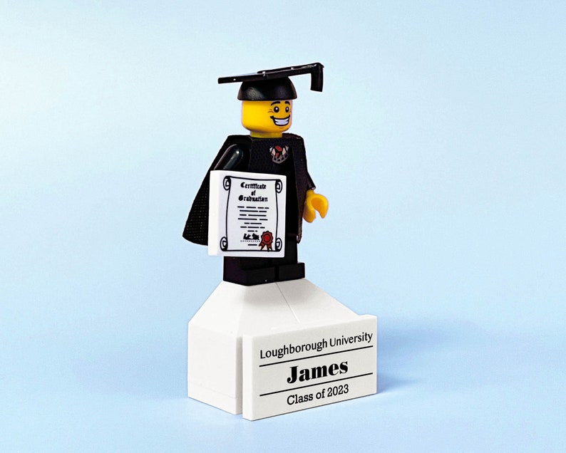 May include: A Lego figure wearing a black graduation gown and cap, holding a certificate. The figure is standing on a white platform with the text "Loughborough University, James, Class of 2023".