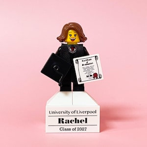 May include: A Lego figurine of a graduate holding a diploma and a mortarboard. The figurine is standing on a white base with the text "University of Liverpool", "Rachel", and "Class of 2027" printed on it. The background is a solid pink.