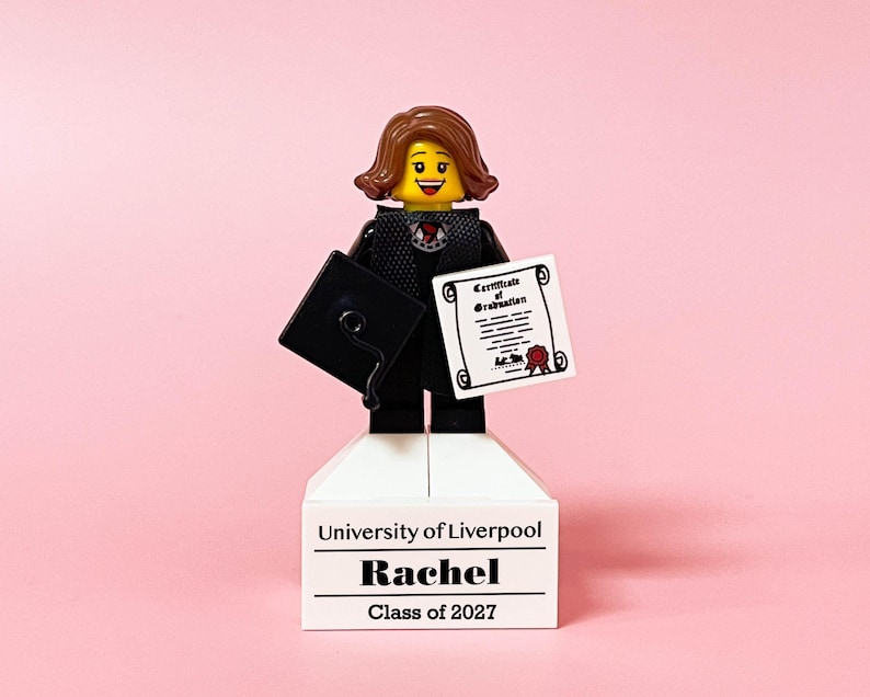 May include: A Lego figurine of a graduate holding a diploma and a mortarboard. The figurine is standing on a white base with the text "University of Liverpool", "Rachel", and "Class of 2027" printed on it. The background is a solid pink.