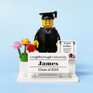 May include: A Lego figure wearing a black graduation gown and cap, holding a diploma. The figure is standing on a white platform with a sign that reads "Loughborough University James Class of 2023". There are three small flowers in a green pot on the platform.