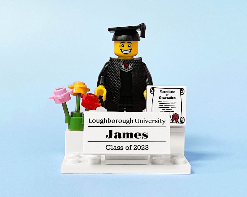 May include: A Lego figure wearing a black graduation gown and cap, holding a diploma. The figure is standing on a white platform with a sign that reads "Loughborough University James Class of 2023". There are three small flowers in a green pot on the platform.