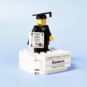 May include: A Lego figure wearing a black graduation gown and cap, holding a certificate. The figure is standing on a white Lego base with a black label that reads "Loughborough University James Class of 2023".