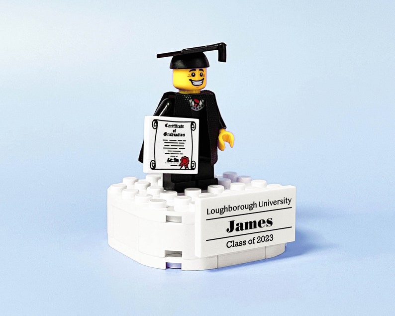 May include: A Lego figure wearing a black graduation gown and cap, holding a certificate. The figure is standing on a white Lego base with a black label that reads "Loughborough University James Class of 2023".