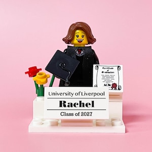 May include: A Lego graduation figure with a black mortarboard, diploma, and a sign that reads "University of Liverpool, Rachel, Class of 2027." The figure is wearing a black graduation gown and has brown hair. Red and yellow flowers are also included.