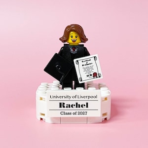 May include: A Lego figurine of a graduate holding a diploma and mortarboard. The figurine is standing on a white base with the text "University of Liverpool", "Rachel", and "Class of 2027" printed on it. The background is pink.