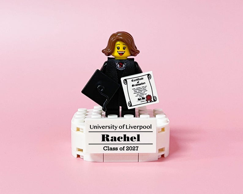 May include: A Lego figurine of a graduate holding a diploma and mortarboard. The figurine is standing on a white base with the text "University of Liverpool", "Rachel", and "Class of 2027" printed on it. The background is pink.