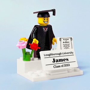 May include: A Lego figurine wearing a black graduation gown and cap, holding a red flower and a diploma. The figurine is standing on a white platform with a sign that reads "Loughborough University, James, Class of 2023".