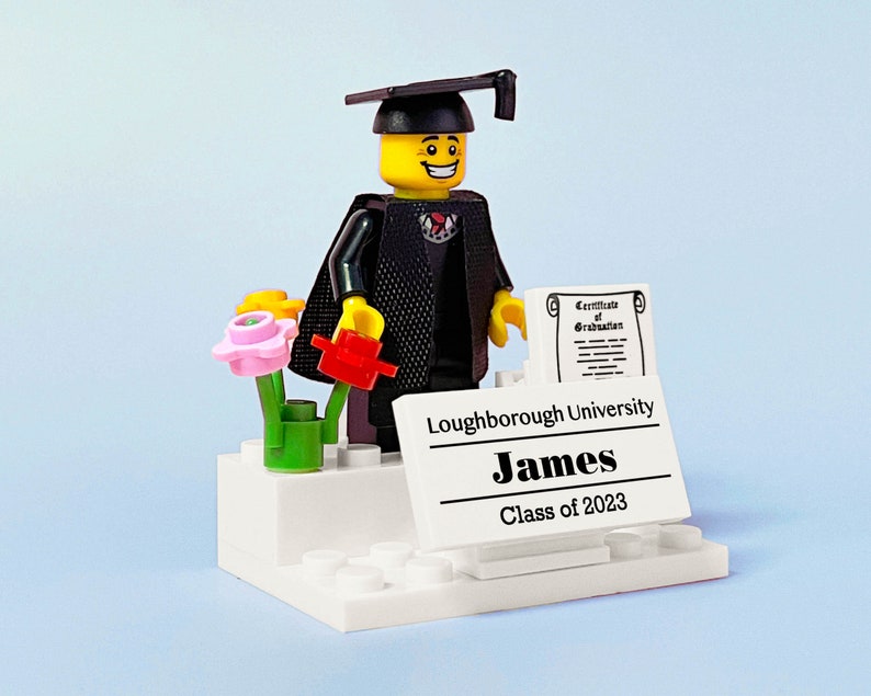 May include: A Lego figurine wearing a black graduation gown and cap, holding a red flower and a diploma. The figurine is standing on a white platform with a sign that reads "Loughborough University, James, Class of 2023".