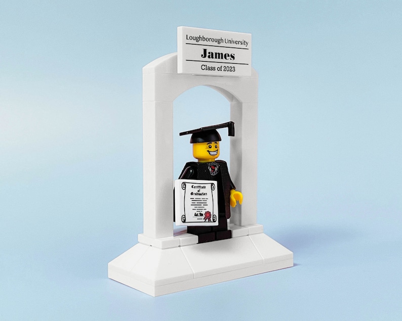 May include: A white LEGO graduation arch with a black-robed LEGO figure holding a diploma. The figure has a yellow face with a smile and a black graduation cap. The arch has a sign that reads "Loughborough University James Class of 2023".