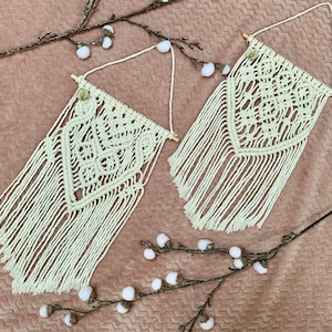 Peut inclure: Deux tentures murales en macramé blanches avec des chevilles en bois et des franges. Les tentures sont réalisées avec une technique de nouage et présentent un motif en losange.