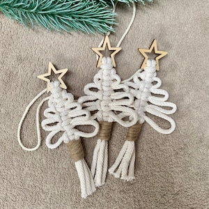 Puede incluir: Tres adornos de árbol de Navidad de macramé blanco con estrellas de madera. Los adornos tienen un borla en la parte inferior y cuelgan de una cuerda blanca.