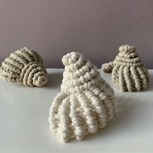 Può includere: Tre conchiglie all'uncinetto in beige e bianco. Le conchiglie sono fatte di corda e hanno forme e dimensioni diverse.