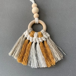 Peut inclure: Un charm de voiture en macramé avec un anneau et des perles en bois. Le charme a un design en forme d'éventail avec des sections alternées de franges blanches, jaune moutarde et grises. Le haut a un anneau en bois et trois perles en bois.