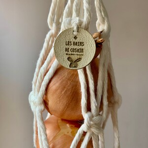 May include: Two brown onions hanging in a white cotton string bag with a leather tag that reads "Les Brins de Coskin" with a rabbit logo.