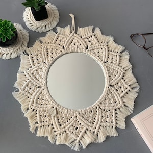 Op de afbeelding: Een witte macramé spiegel met een ronde spiegel in het midden. De spiegel is omgeven door een wit macramé ontwerp met franjes aan de randen.