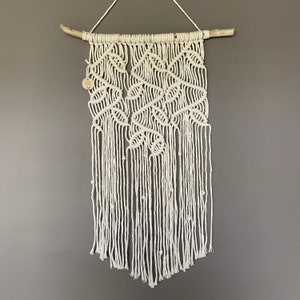 Puede incluir: Un tapiz de macramé blanco con un patrón geométrico, que presenta una rama de madera natural en la parte superior.