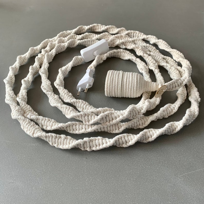 Op de afbeelding: Cr&egrave;mekleurig macram&eacute; snoer met een witte in-line schakelaar, stekker en lamphouder. Het snoer is opgerold en heeft een getextureerd, gedraaid ontwerp. Het snoer is ontworpen voor gebruik met een lamp.