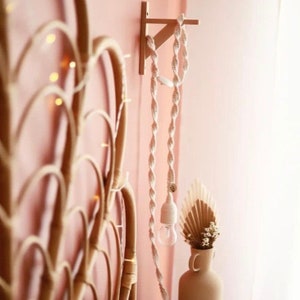Op de afbeelding: Een cr&egrave;mekleurige macram&eacute; hanglamp met een spiraalvormig ontwerp hangt aan een houten beugel. Een vaas met gedroogde bloemen staat onder de lamp. Een rotan hoofdbord is zichtbaar op de achtergrond. De muur is roze.