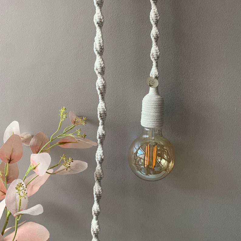Op de afbeelding: Een macram&eacute; hanglamp met een grote, ronde Edison-lamp. Het licht wordt opgehangen door een geweven, gebroken wit touw. De lamp is omhuld in een bijpassende geweven kap. Een takje roze bloemen staat in de linker benedenhoek.