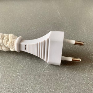 Op de afbeelding: Close-up van een witte stekker met twee zilverkleurige pinnen. De stekker is verbonden met een cr&egrave;mekleurig, gevlochten snoer. De stekker heeft een geribbeld ontwerp aan de zijkant.