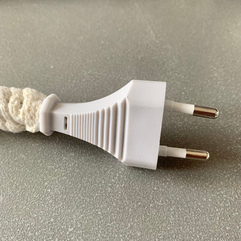 Op de afbeelding: Close-up van een witte stekker met twee zilverkleurige pinnen. De stekker is verbonden met een cr&egrave;mekleurig, gevlochten snoer. De stekker heeft een geribbeld ontwerp aan de zijkant.